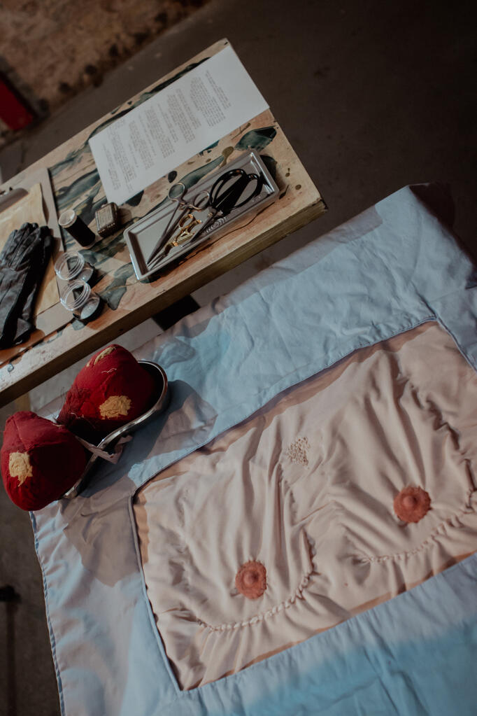 A quilt that looks like a human chest with a blue medical sheet is sat on a table. There is a small metal dish with fabric breast tissue sat on the edge of the quilt. Above this is another metal dish, this one filled with an assortment of medical tools.