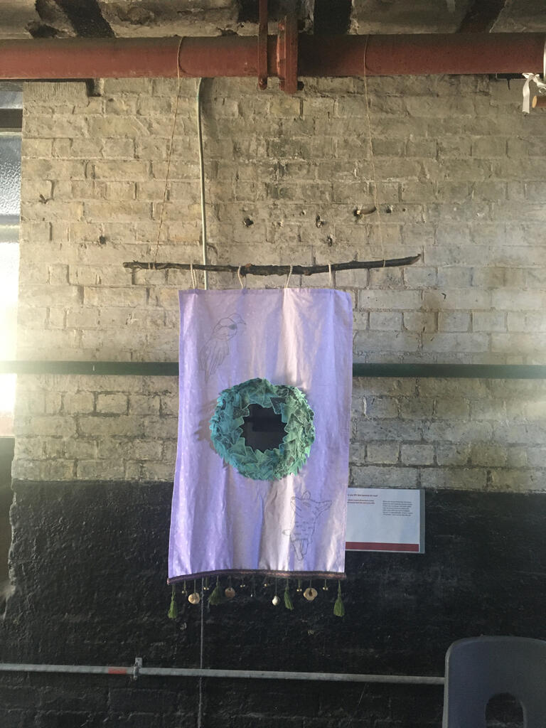 A purple wall hanging, featuring silver prints of a magpie and a fox. In the centre is a gloss black disc surrounded by fabric ivy leaves. The bottom features a woven border with tassels, bells and hags stones. This is hung from a pipe above it, and is set